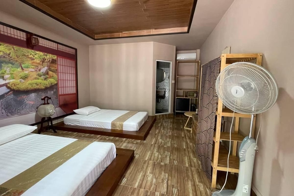 Habitación de hotel con dos camas, suelo de madera, un ventilador grande y una pintura mural tradicional al estilo japonés.