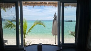 A room with a large window overlooking a beach with clear turquoise water, a palm tree, and a thatched roof structure. - White Sand Ark Resort (Koh Rong)