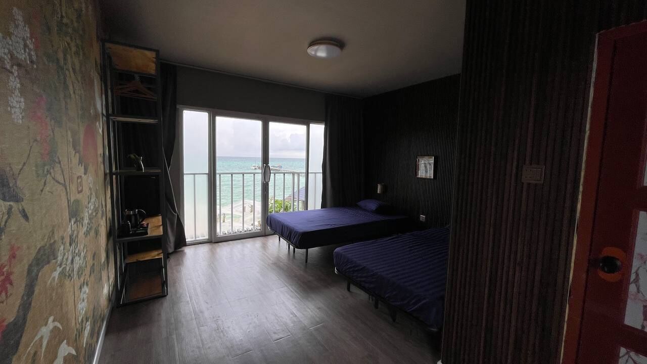 A room with two beds, a large window with a view of the sea, and a sliding door leading to an outdoor balcony.