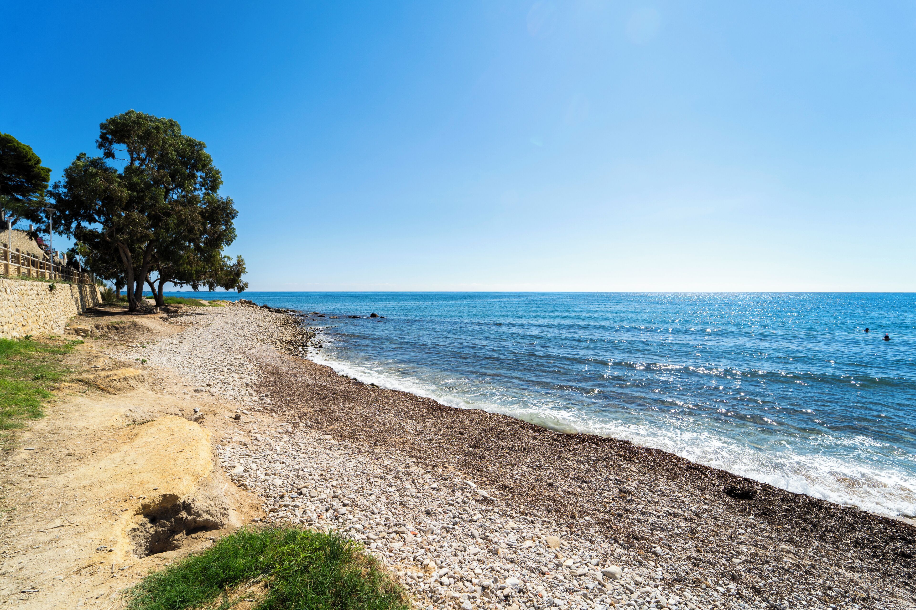 Une plage de galets bordée d’un muret et d’arbres, qui mène à la mer.