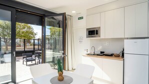 Cocina moderna con armarios blancos, microondas y refrigerador. Hay una zona de comedor con mesa y sillas, y un puerta de cristal que da a una terraza.