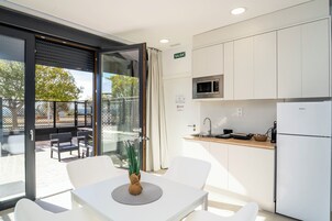 Cocina moderna con armarios blancos, microondas y refrigerador. Hay una zona de comedor con mesa y sillas, y un puerta de cristal que da a una terraza.