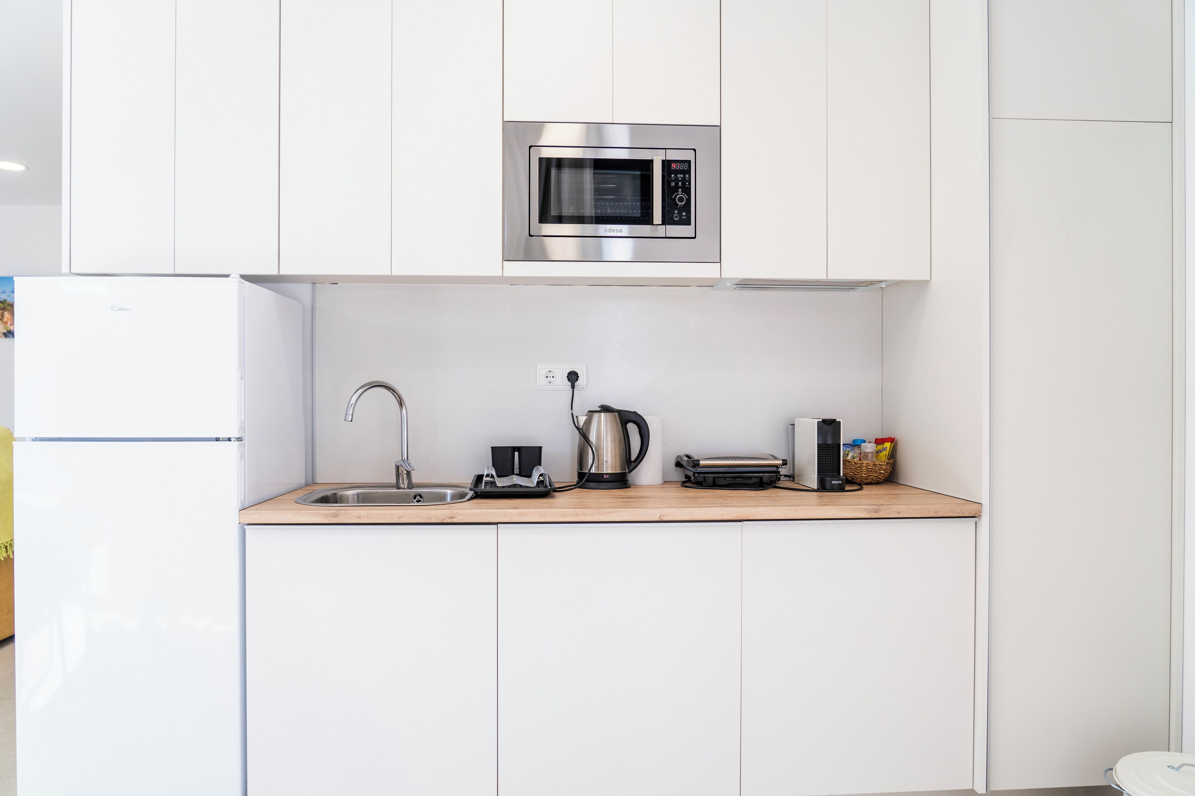 A modern kitchen with white cabinets, a stainless steel microwave, a kettle, a toaster, and a sink.