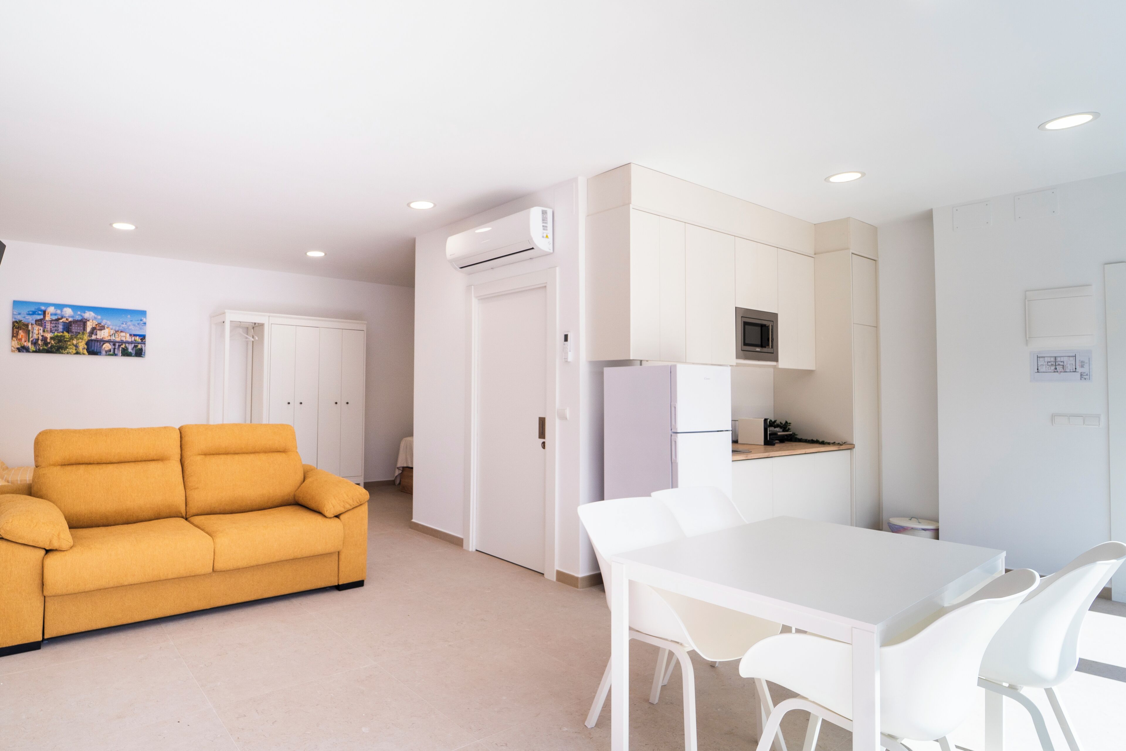 A modern kitchen with white cabinets, a microwave, and a refrigerator. There is a white dining table with chairs. A mustard-colored sofa is in the living area.