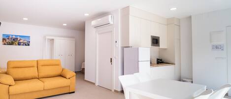 A modern kitchen with white cabinets, a microwave, and a refrigerator. There is a white dining table with chairs. A mustard-colored sofa is in the living area.