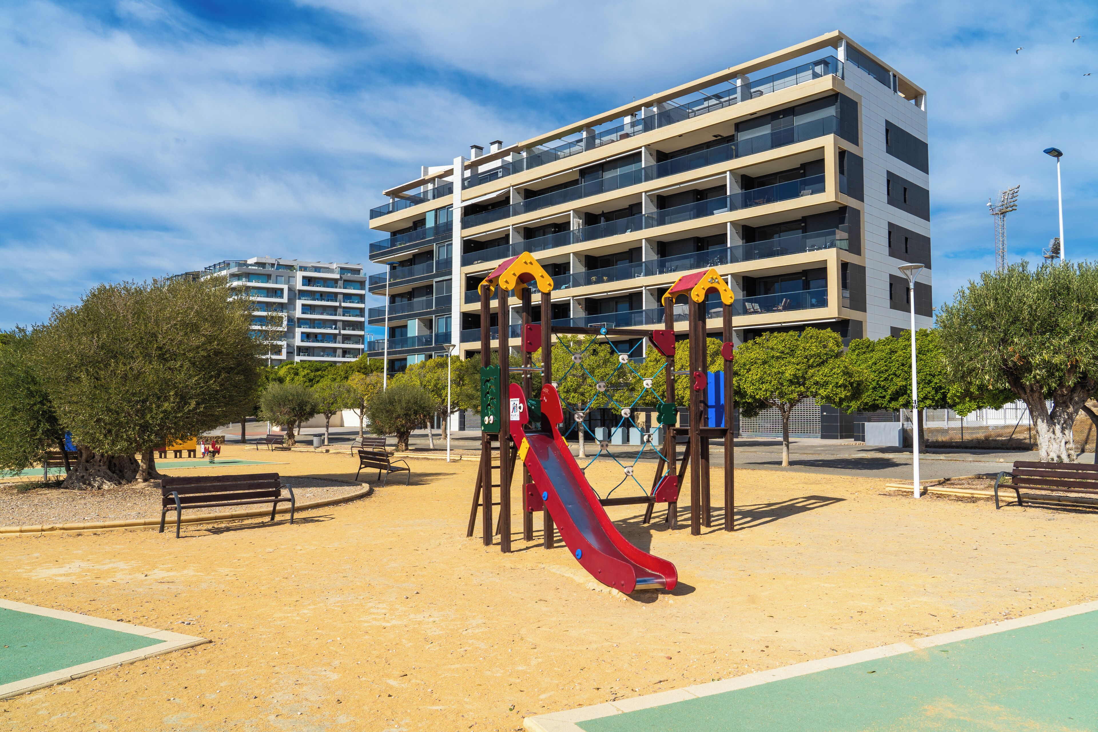Un terrain de jeux avec un toboggan et des balançoires, des bancs et un bâtiment en arrière-plan.