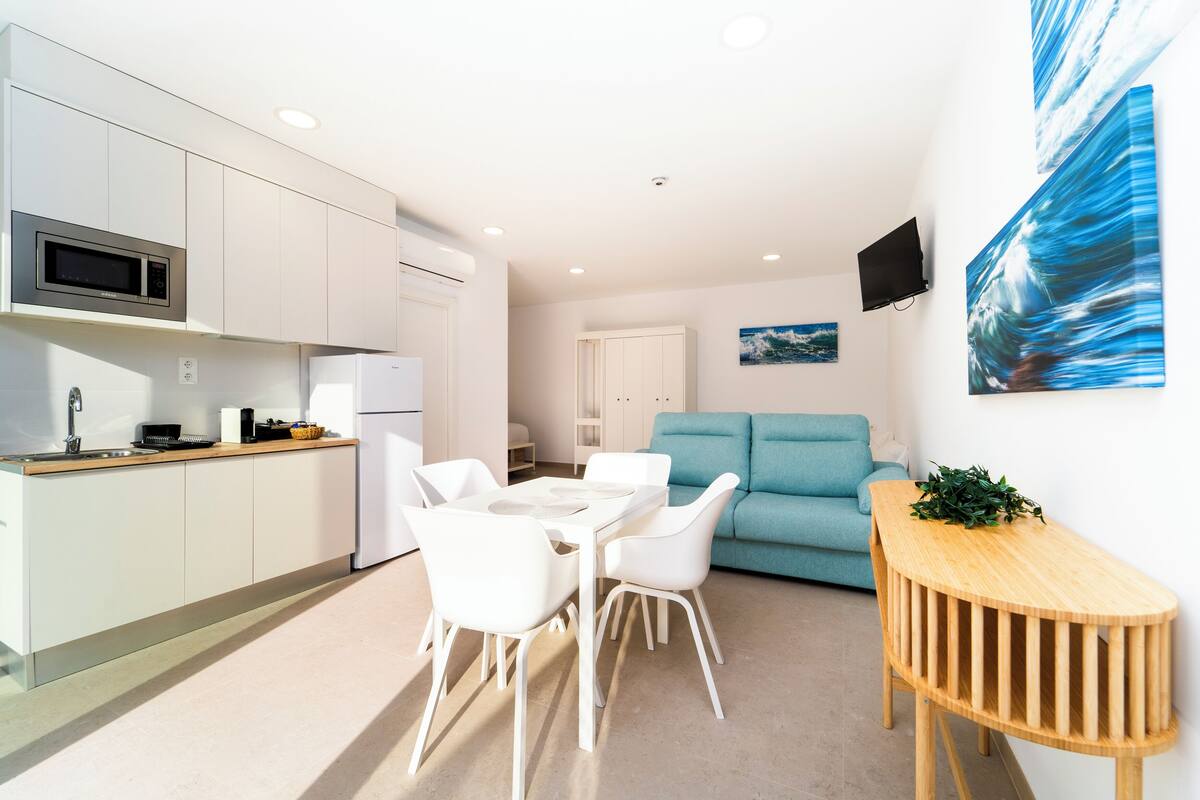 Une cuisine moderne avec des meubles blancs, un four à micro-ondes et un réfrigérateur. On y trouve un coin repas avec une table et des chaises blanches. Un canapé de couleur turquoise se trouve dans le salon, et une petite table en bois est placée à côté.