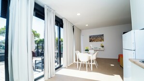 Una cocina moderna con electrodomésticos blancos, un comedor con mesa y sillas, y ventanas grandes con cortinas.