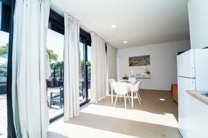 Una cocina moderna con electrodomésticos blancos, un comedor con mesa y sillas, y ventanas grandes con cortinas.