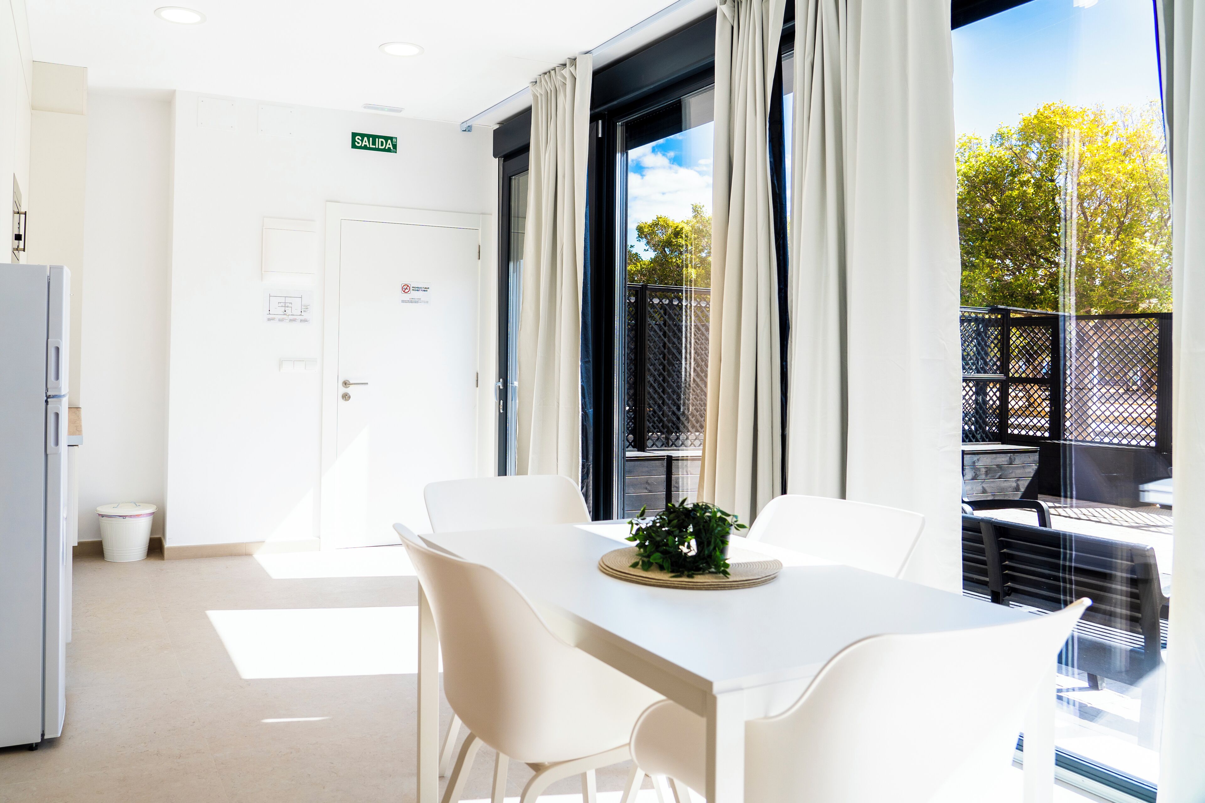 A bright room with a white table and chairs, a door with a sign, and a view of an outdoor area with a fence.
