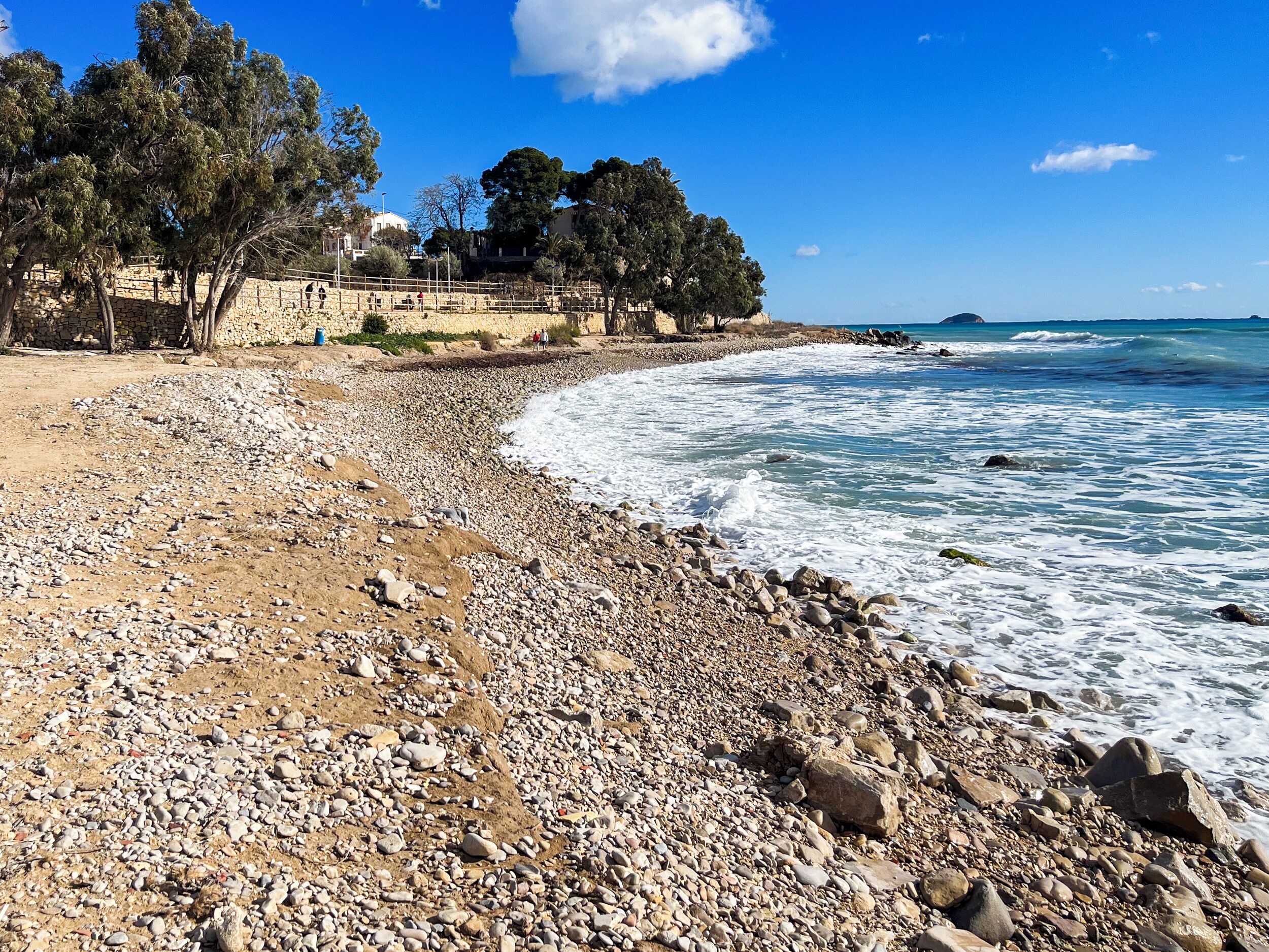 A pebble beach with a stone wall and trees, leading to a rocky shoreline.