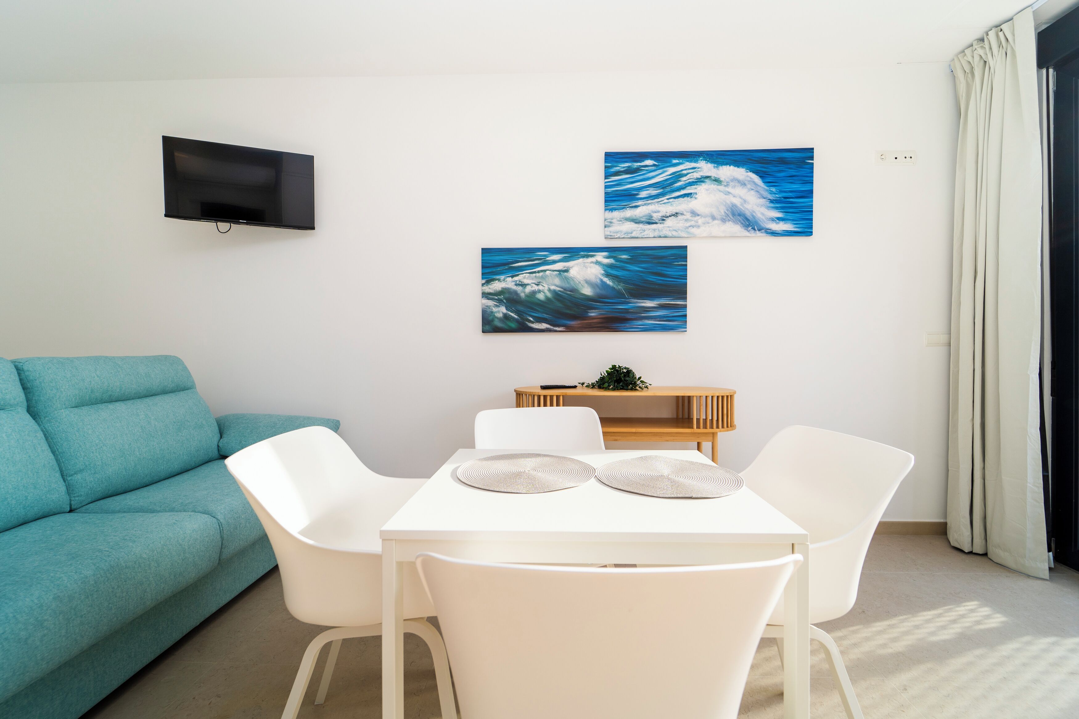 A modern living room with a teal sofa, a white dining table with two chairs, and two wall-mounted paintings of ocean waves.