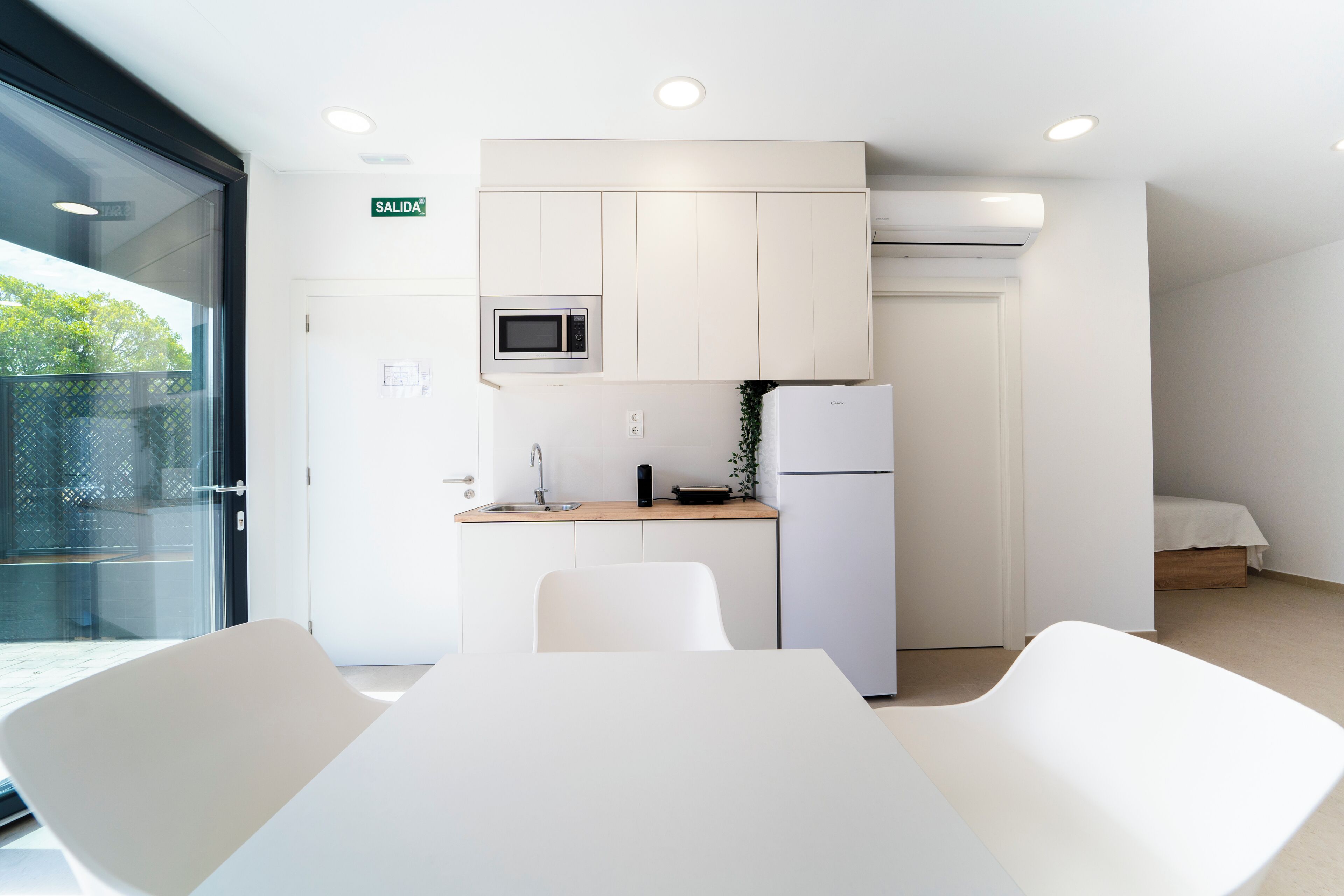 A modern kitchen with white cabinets, a microwave, and a refrigerator. There is a dining area with white chairs and a table. A door labeled "SALIDA" is visible, indicating an exit.