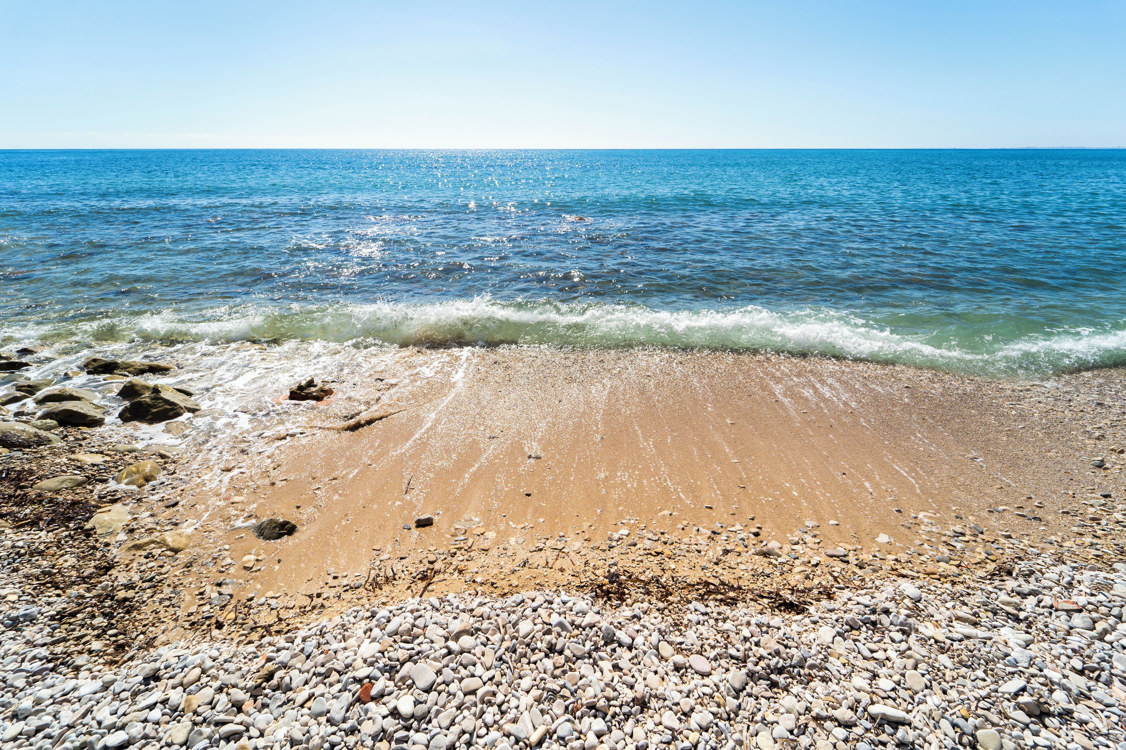 A beach with pebbles and sand, a clear blue sea, and a calm horizon.