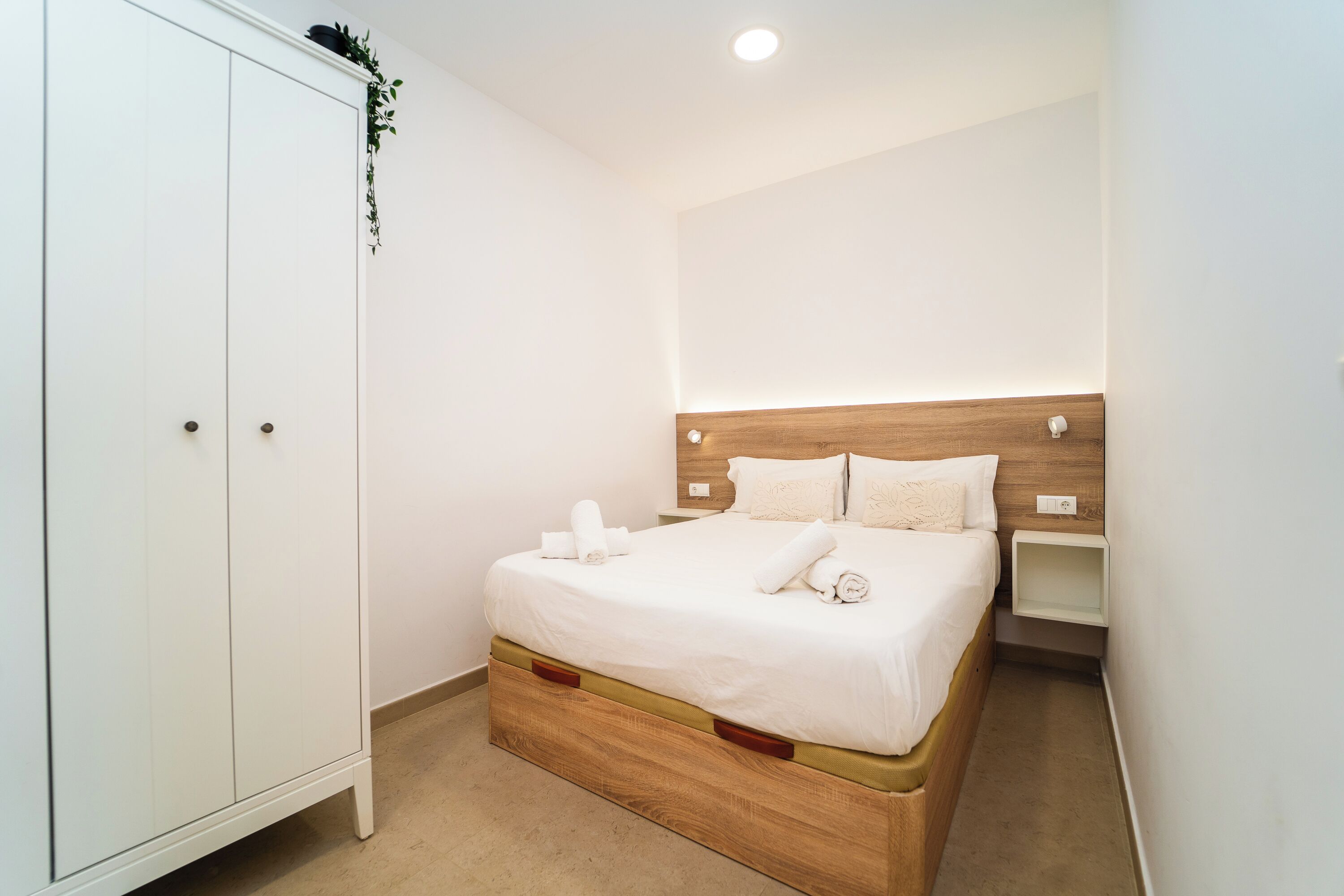 A bedroom with a wooden headboard, a white wardrobe, and a bedside table.
