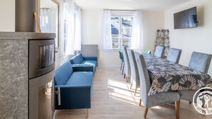 Ein modernes Wohnzimmer mit Sofa, Esstisch und Stühlen. Ein Kamin, ein an der Wand befestigter Fernseher und große Fenster mit Vorhängen sind ebenfalls vorhanden.