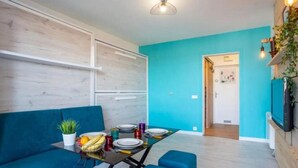 A modern living space with a teal wall, a blue sofa, a dining table with plates and fruits, and a TV mounted on the wall. - Charming studio for 4 in Ciboure with terrace (Ciboure)