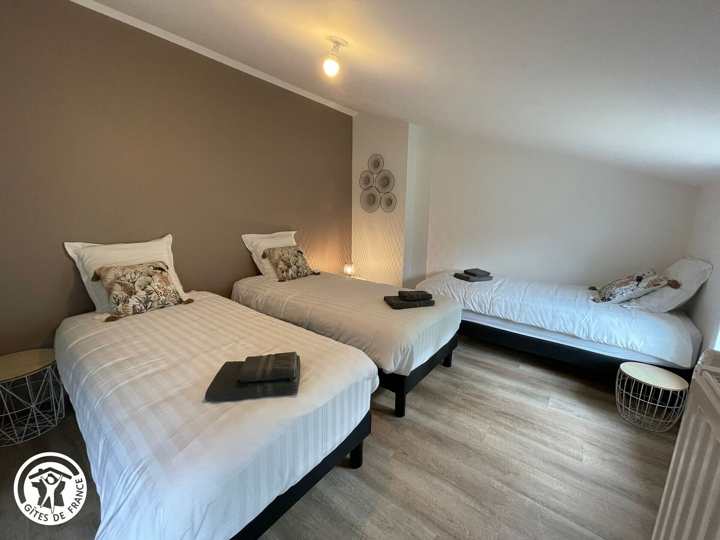 Hotellihuoneessa on kolme erillistä sänkyä, jokaisessa on tyyny ja taitettu pyyhe. Sänkyjen yläpuolella on pieni pyöreä pöytä ja koristeellinen seinäkoriste.