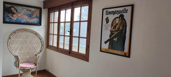 Una habitación con una silla de mimbre, un ventanal grande y dos carteles enmarcados en la pared.