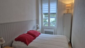 Une chambre à coucher comprenant un lit, une table de chevet, une lampe, une fenêtre avec des stores et un radiateur.