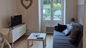 Un salon avec un téléviseur posé sur un meuble blanc, une décoration en forme de cœur accrochée au mur, et une vue sur la verdure à l’extérieur, visible à travers une fenêtre donnant sur un balcon.