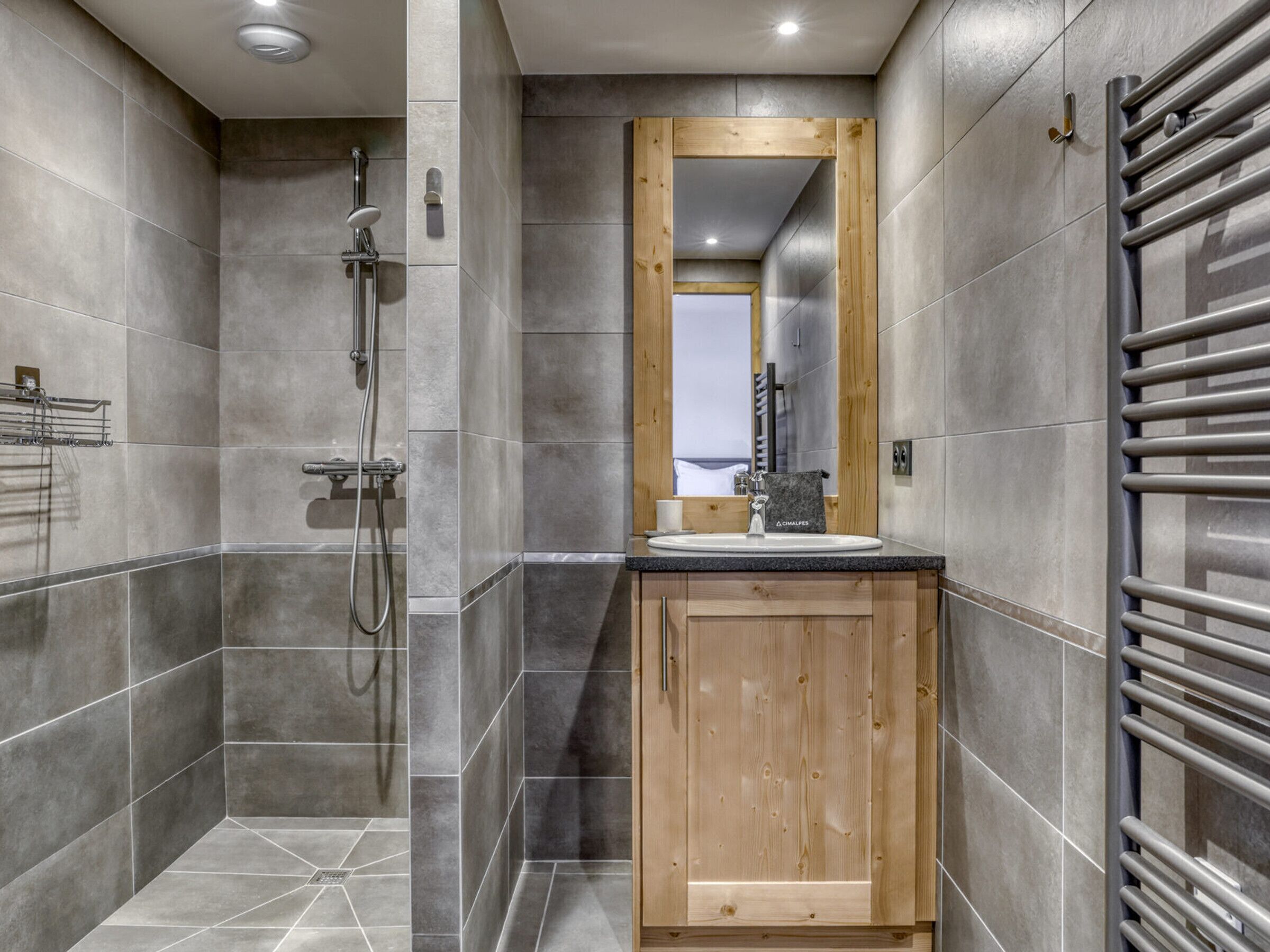 A modern bathroom with grey tiles, a wooden cabinet, a sink, a mirror, and a shower.