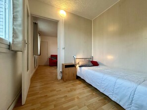 Un petit appartement d’une seule pièce, avec un plancher en bois, un lit recouvert de draps blancs, une chaise rouge et un petit meuble en bois.
