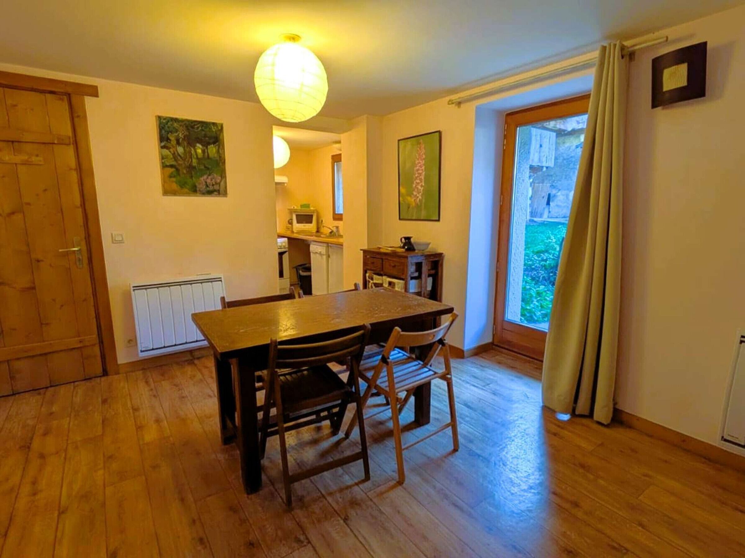 Ein Essbereich mit Holztisch und Stühlen, im Hintergrund eine Küche und durch ein großes Fenster mit Vorhängen ein Ausblick.