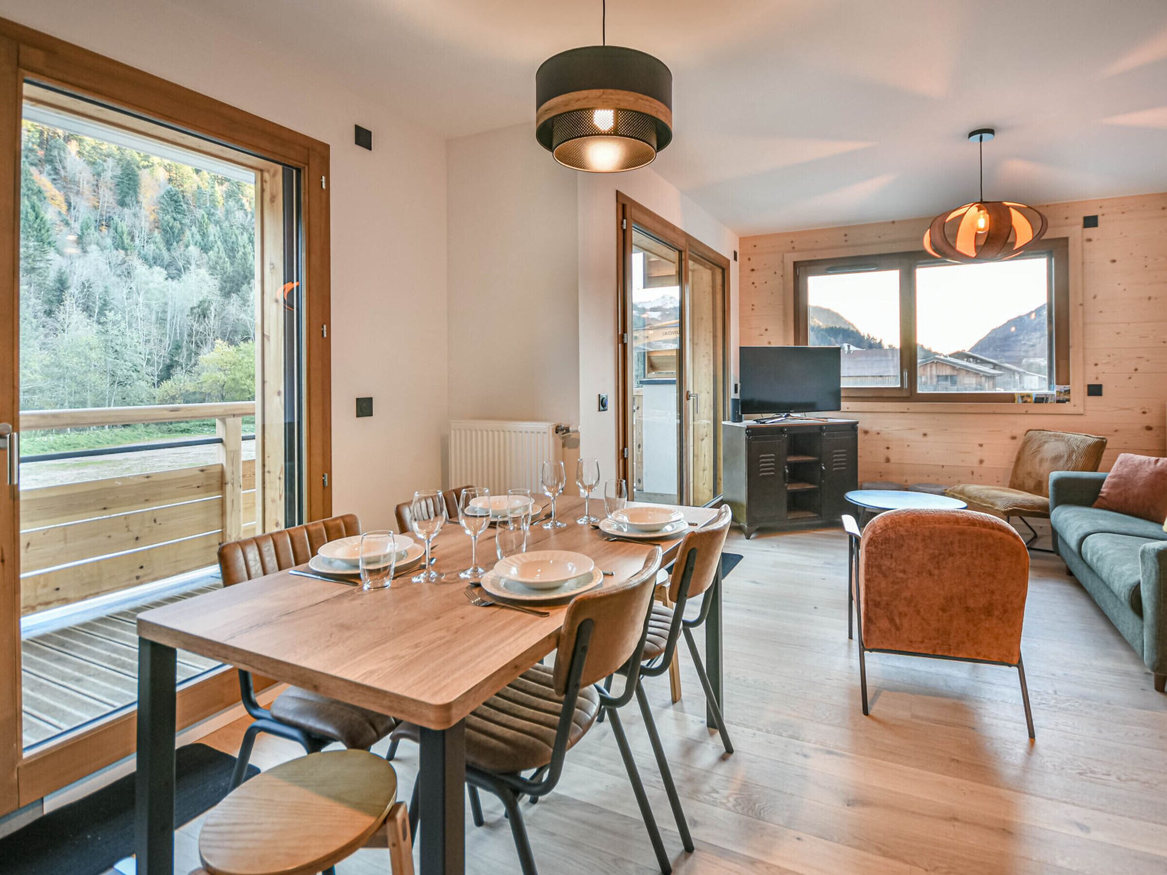 A modern, well-lit dining area with a wooden table set for six, surrounded by chairs. A television is mounted on the wall, and there is a sofa and a chair in the adjacent living area.