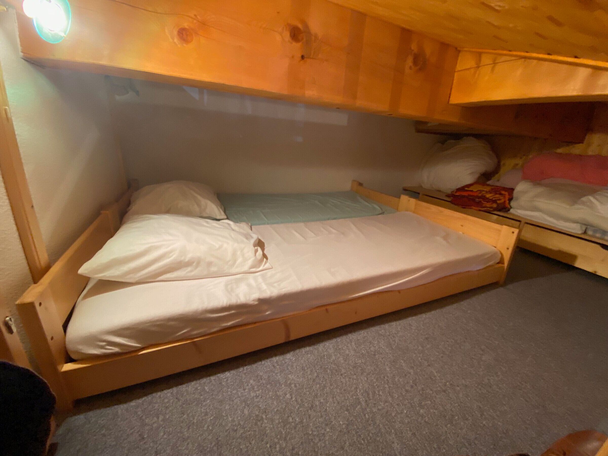 A small, single-bed room with a wooden bunk bed structure, a carpeted floor, and a ceiling light fixture.