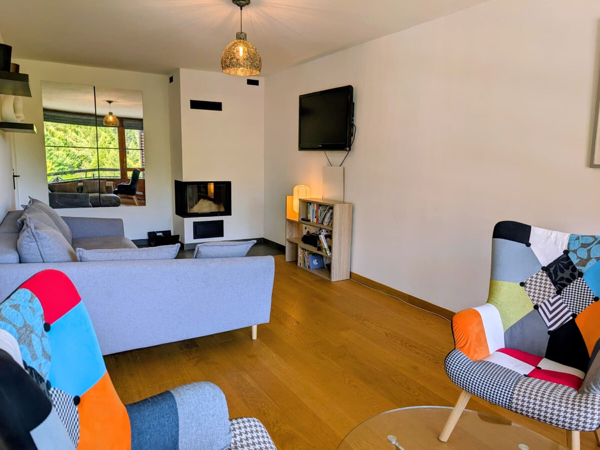 A modern living room with a grey sofa, colorful armchairs, a flat-screen TV, and a bookshelf.