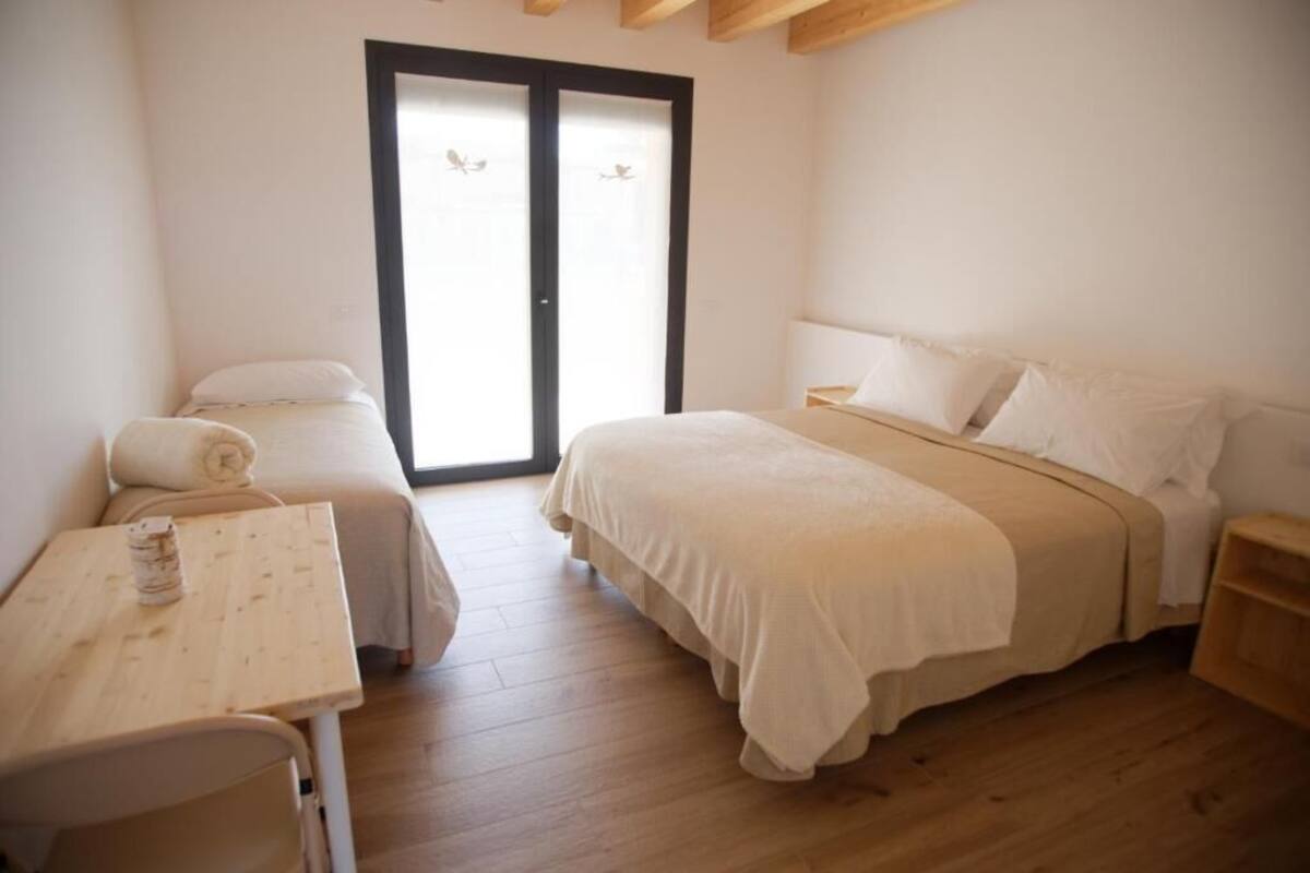 Een slaapkamer met een bed, een klein tafeltje en een stoel. Er is een schuifpui die uitkomt op een buitengebied.