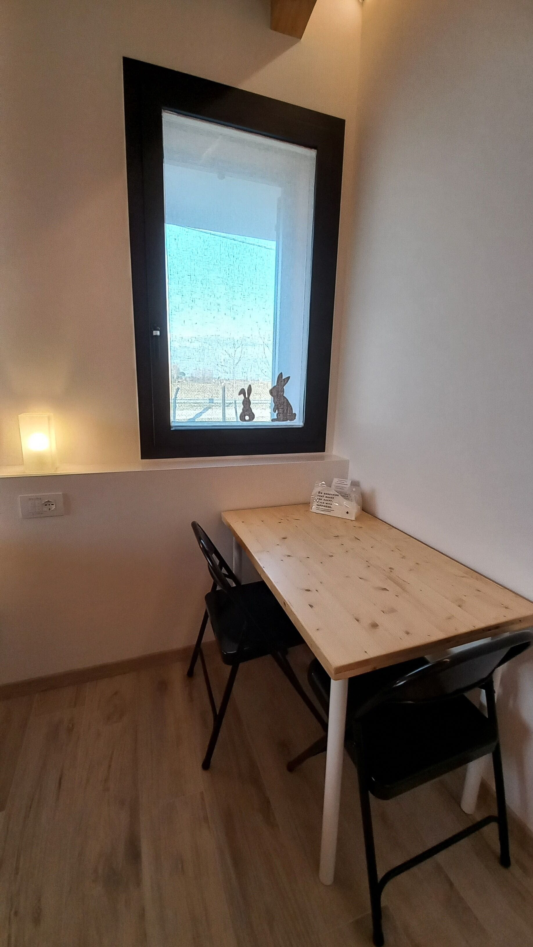 Una habitación pequeña con una mesa de madera y dos sillas, una ventana que ofrece vista a un edificio, y un enchufe de pared.