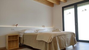 Een slaapkamer met een houten vloer, een bed met een beige deken, een nachtkastje, een houten plank en een raam met uitzicht op vogels.