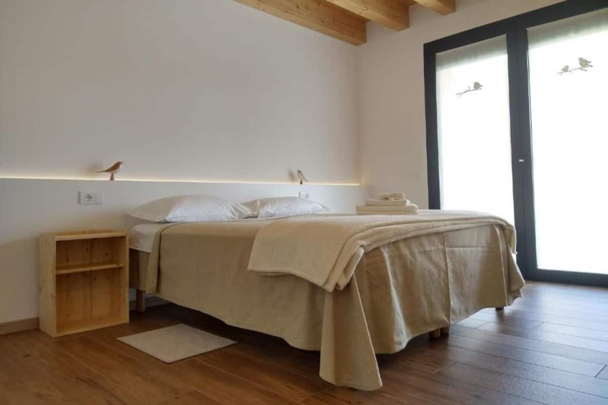 Een slaapkamer met een houten vloer, een bed met een beige deken, een nachtkastje, een houten plank en een raam met uitzicht op vogels.