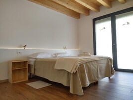 Un dormitorio con piso de madera, una cama con manta color beige, una mesita de noche, un estante de madera y una ventana con vista a pájaros.