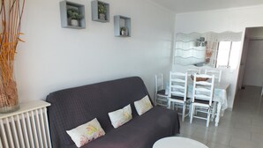 Una sala moderna con un sofá gris oscuro, una mesa de centro redonda blanca y un comedor con sillas blancas y una mesa con superficie de vidrio.