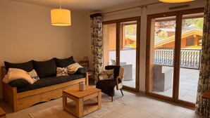 A cozy living room with a sofa, a coffee table, and a chair. There is a large window with curtains, and a view of a building outside. - Cozy apartment with large terrace and ski locker (Pralognan-la-Vanoise)