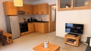 A compact kitchen with wooden cabinets, a built-in oven, and a sink. There's a small wooden table with a kettle, a TV on a stand, and a sofa in the living area. - Cozy apartment with large terrace and ski locker (Pralognan-la-Vanoise)
