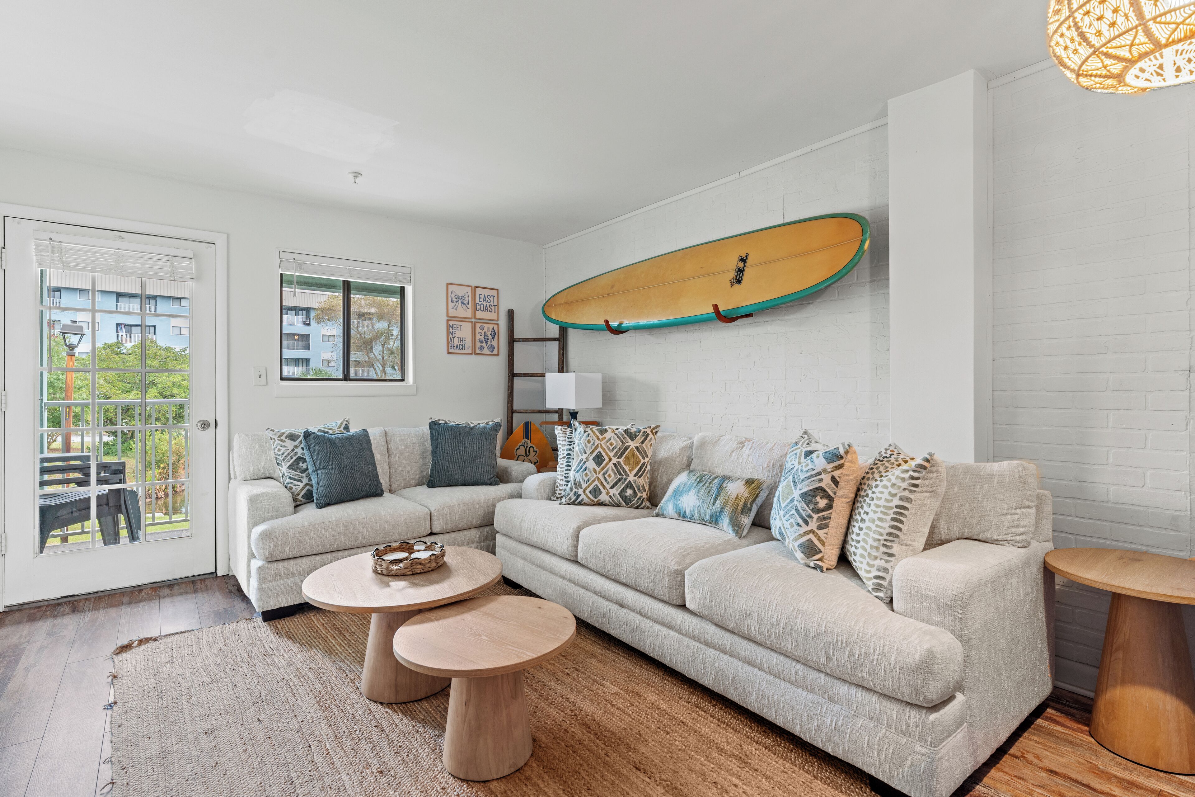 Ein Wohnzimmer mit einem Ecksofa, zwei runden Holztischen, einem Surfbrett an der Wand und Blick auf einen Balkon mit Gartenmöbeln.