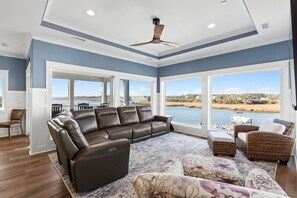 A spacious living room with a large window, a ceiling fan, and a leather sofa. - Docks and Dunes: A spacious 5-bedroom beach house in Oak Island, perfect for 14 guests! (Oak Island)