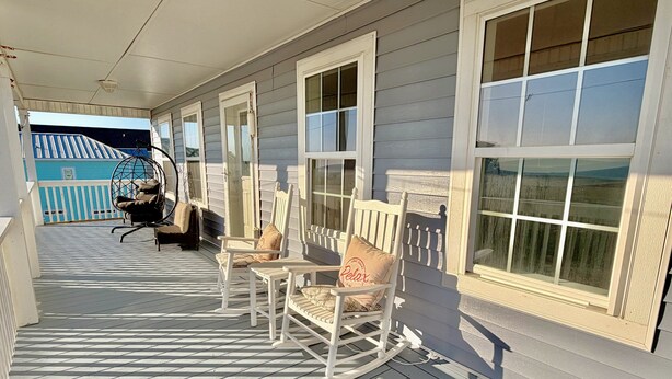 A covered porch with rocking chairs, a hanging chair, and a view of a blue building. - Oceanview retreat in Oak Island, sleeps 12, perfect for families and a dog! (Oak Island)