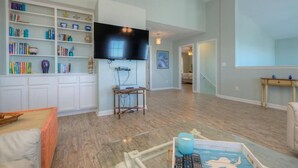 A spacious living room with a flat-screen TV mounted on the wall, a bookshelf filled with books and decorative items, and a wooden floor. - Oceanview retreat in Oak Island, sleeps 12, perfect for families and a dog! (Oak Island)