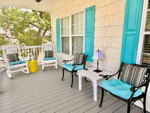 A covered porch with wooden flooring, white rocking chairs with teal cushions, a small table with a lamp, and turquoise shutters on the windows. - Nemo's Place: Spacious 4BR retreat with private pool in scenic Oak Island. (Oak Island)
