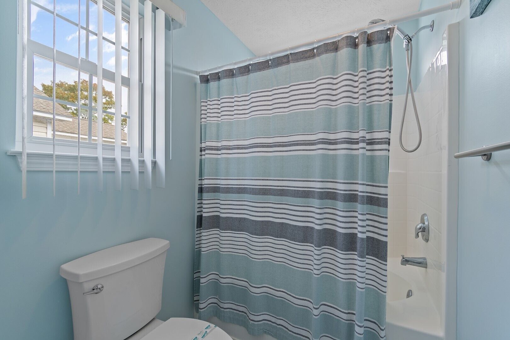 A bathroom with a toilet, a shower with a striped curtain, and a window with blinds.