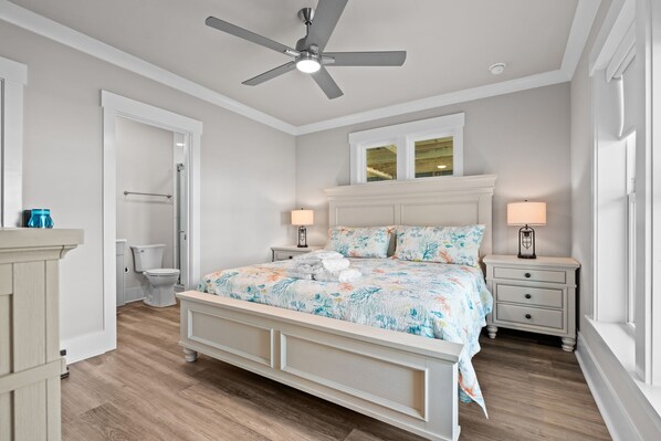 Quarto com cama, ventilador de teto, janela, mesa de cabeceira e banheiro com vaso sanitário e chuveiro.