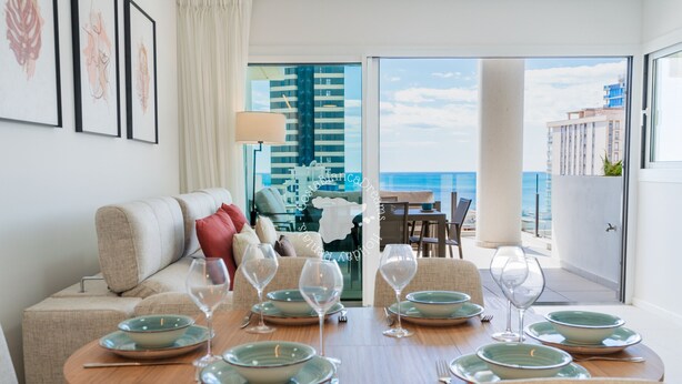 Ein Essbereich mit Blick auf das Meer und Gebäude, eine Couch mit Kissen sowie ein Tisch gedeckt mit Tellern und Weingläsern.