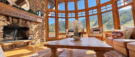 A cozy living room with a stone fireplace, wooden furniture, and large windows offering a view of the outdoors.