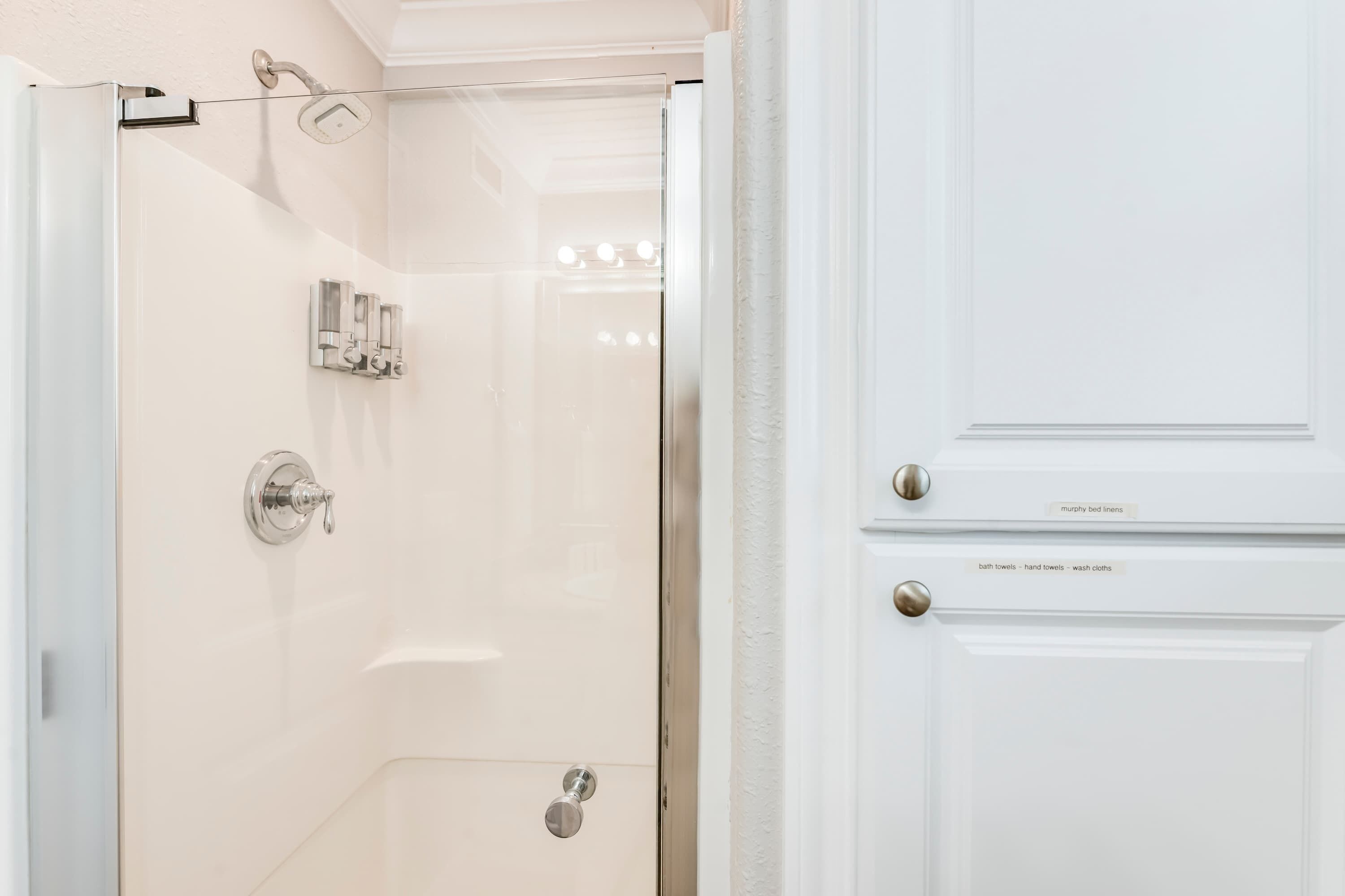 A modern bathroom with a glass shower enclosure, a white vanity, and a door labeled "murphy bed linens."