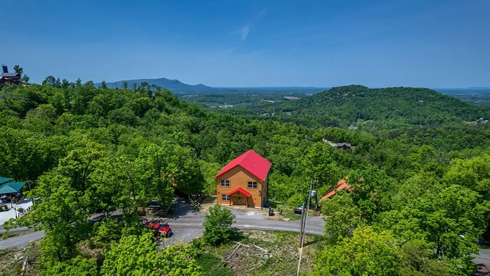 A house with a red roof surrounded by dense green forest, with a clear sky and distant mountains.